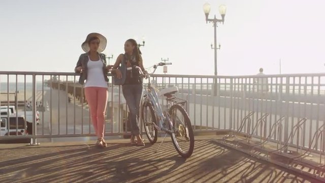 Two black women best friends chatting and leaning against metal railing at the pier with the sun behind them