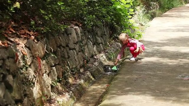 Girl running along the path with a paper boat in hand. She stops near the creek and runs it in the swimming
