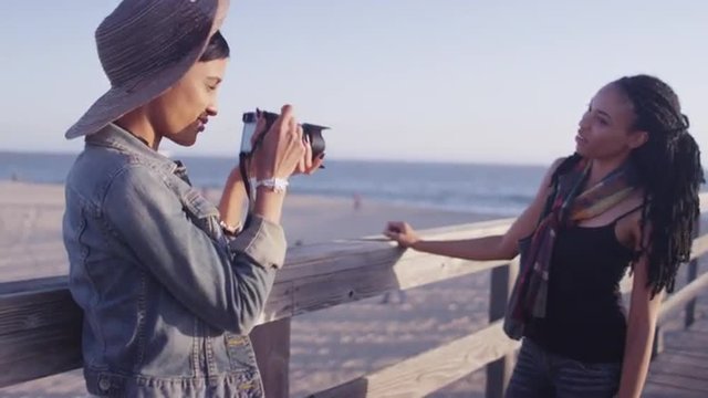 Black women best friends taking pictures of each other with camera on the pier