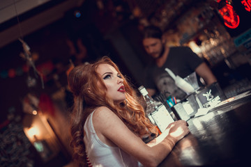 Sad sexy woman sitting on the bar counter at night with glass of whiskey
