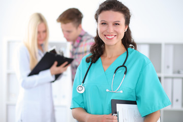 Fototapeta premium Beautiful young smiling female doctor standing at hospital with doctor and patient on the background. Medical concept.