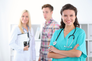 Fototapeta premium Beautiful young smiling female doctor standing at hospital with doctor and patient on the background. Medical concept.