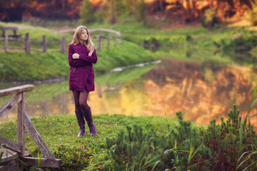 Outdoor photo from a beautiful young women standing before small lake in a park, with a violet colored clothing at autumn