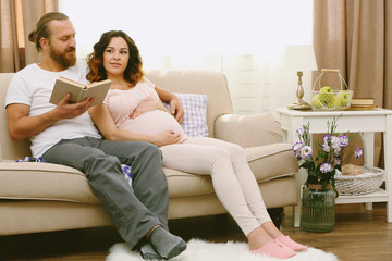Fototapeta na wymiar Handsome man reads book to his lovely pregnant woman on sofa in the room