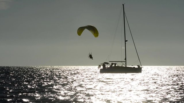 Motorized paraglider low pass near a sailboat at sunset on the sea