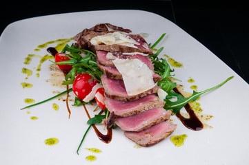 Medium rare steak served on a plate with cherry tomatoes and arugula. On top of it is grated parmesan cheese.