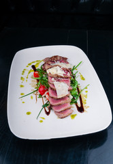 Medium rare steak served on a plate with cherry tomatoes and arugula. On top of it is grated parmesan cheese.