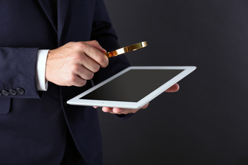 Businessman holding magnifying glass and digital tablet on dark grey background, searching and examining concept