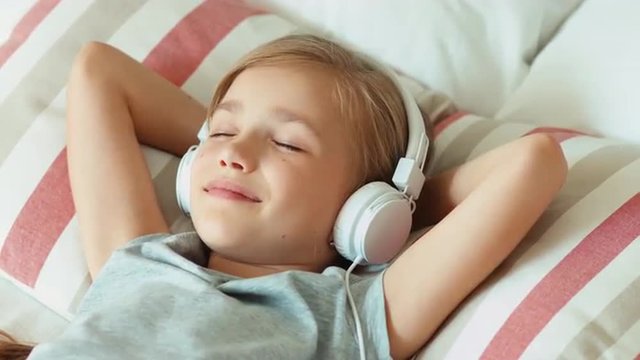 Closeup portrait girl child listening music in headphones with eyes closed and lying on the bed and resting and smiling at camera