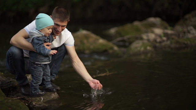 Father with little son playing on the river bank with water. Warm autumn. Daddy shows son of water in the river. Son sees dad squelching water. 
