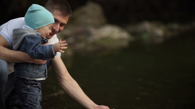 Father with little son playing on the river bank with water. Warm autumn. Daddy shows son of water in the river. Son sees dad squelching water. 