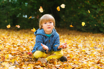 Autumn portrait of a cute little boy of 4 years old, playing with yellow leaves in the park, wearing blue jacket and yellow trousers