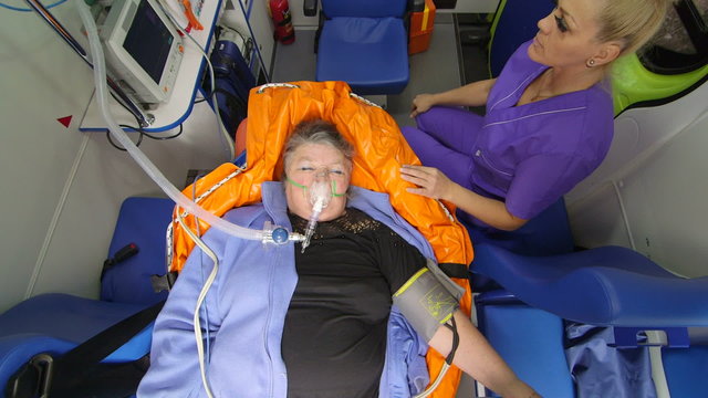 Female EMT paramedic provide medical care to critical senior woman patient with eyes closed in ambulance, camera directly above
