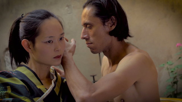 A Japanese woman in a traditional geisha outfit and a shirtless Mexican man performing as butoh modern dance dancers with slow hyper controlled movements.