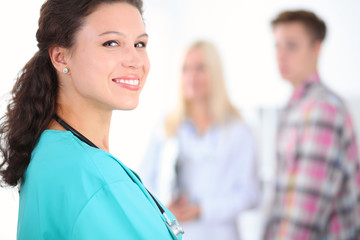 Beautiful young smiling female doctor standing at hospital with doctor and patient on the background. Medical concept.