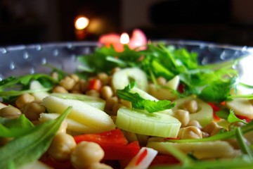 Fresh green salad including cucumber, apple, chickpea, paprika and arugula served on a glass bowl with candle on the background. 