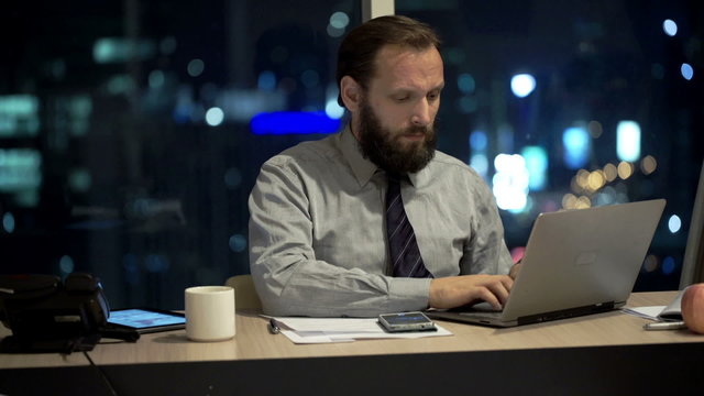 Angry businessman throwing laptop in office at night
