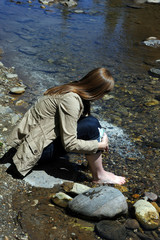 Young woman sits barefoot besides the Gallatin River in the Gallatin Valley of Montana.  She is picking up rocks from the riverbed.