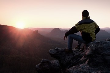 Rear view of male hiker in yellow black jacket sitting on rocky cliff while enjoying daybreak above...