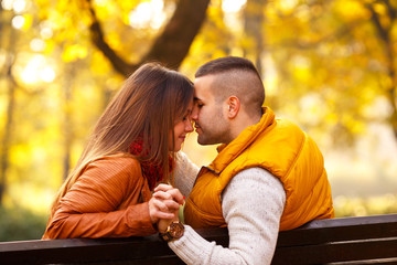 Fototapeta premium Young couple sitting on a park bench on a sunny autumn day and expressing tenderness