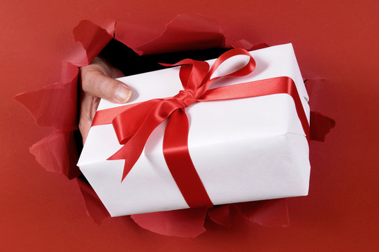 Christmas Or Birthday Gift Package Parcel Being Delivered Or Bursting Held By Man Hands Through Torn Red Paper Background Shipping Delivering Giving Present Photo