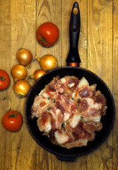  Fresh raw meat in a frying pan with onion and tomatos near it. Wooden background.
