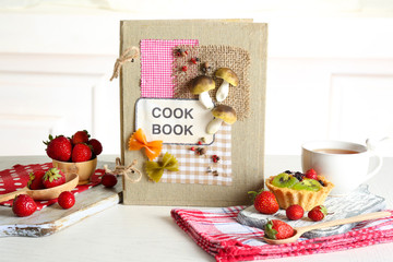 Decorated cookbook with cup of tea and tasty cake on white background