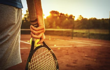 Foto op Aluminium Man holding tennis racket/Close up of man holding tennis racket on clay court. In his hand is tennis ball. On court is sunset. © likoper