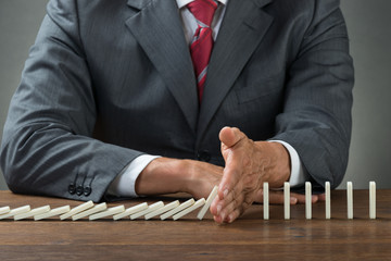 Businessman Stopping Dominoes Falling On Wooden Desk
