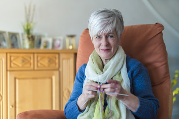 Eine Seniorin sitzt mit Strickzeug in ihrem Sessel im Wohnzimmer.
