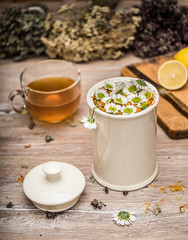 Herbal tea. Medicative herbs in a ceramic can and in bunches, tea in a glass cup, a lemon and a knife on a chopping board.