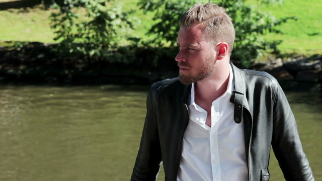 A man wearing a black leather jacket and white shirt, relaxing outside with the river behind him on a sunny summer day.