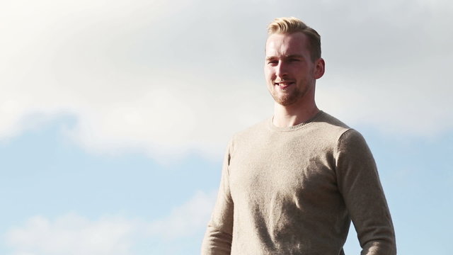 A lonely man standing against a blue sky wearing a warm pullover shirt smiling against camera on a sunny day.