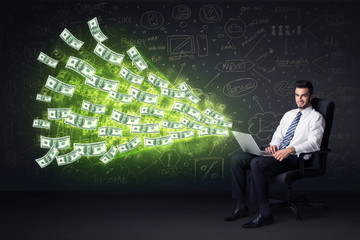 Businessman sitting in chair holding laptop with dollar bills co