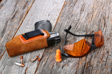 Automatic Handgun with leather holster, bullets and safety glasses on a wooden background.