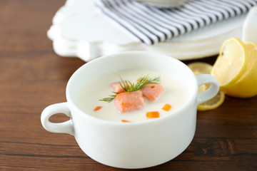 Delicious salmon cream soup in white bowl with lemon and striped cotton serviette on wooden table, close up