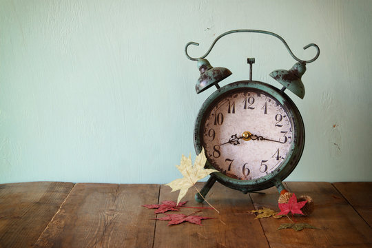 image of vintage alarm clock next to autumn leaves on wooden table in front of wooden background. retro filtered
