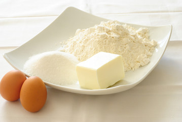 ingredienti per preparare torte, biscotti, cupcakes, in un piatto quadrato ci sono un monte di farina, un monte di zucchero, un pezzo di burro, e appoggiati al piatto due uova