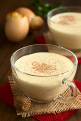 Eggnog drink in glass cups sprinkled with ground nutmeg, eggnog cake baked in eggshell in the back, photographed on dark wood with natural light (Selective Focus, Focus  in the middle of the eggnog)