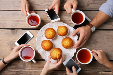 Four hands with smart phones holding  cups with tea, on wooden table background