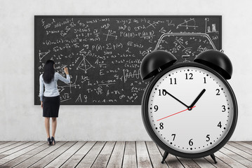 Rear view of business lady who is writing math formulas on the black chalkboard. The huge alarm clock is on the foreground. A concept of time and quantitative science. Wooden floor and concrete wall.