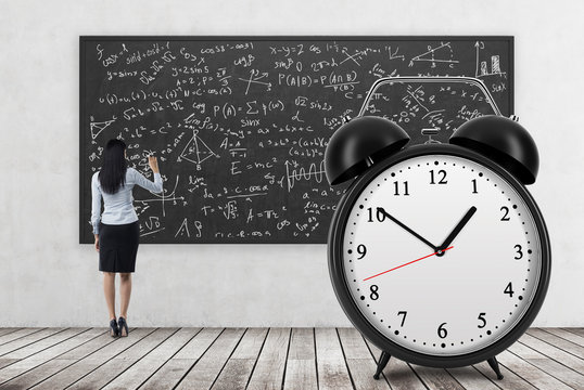 Rear view of business lady who is writing math formulas on the black chalkboard. The huge alarm clock is on the foreground. A concept of time and quantitative science. Wooden floor and concrete wall.