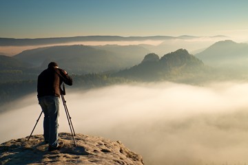 Professional on cliff. Nature photographer takes photos with mirror camera on rock. Dreamy fogy landscape, spring orange pink mist below.