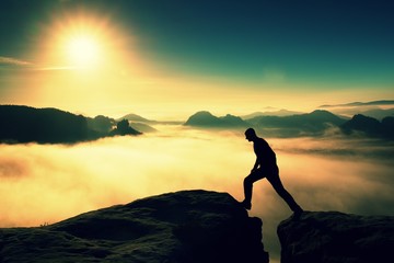 Fototapeta premium Crazy hiker in black is jumping between the rocky peaks. Wonderful daybreak in rocky mountains, heavy mist in deep valley. Miracle of nature