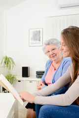 cheerful young woman reading book for old senior woman at home