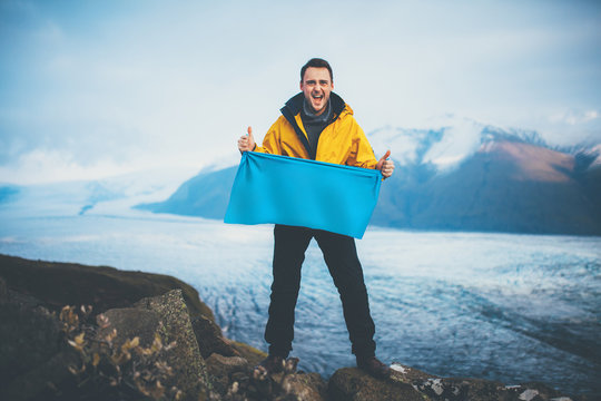 Smiling young man holding a blank flag on top of mountain. Male wearing winter wear holding blue empty banner at glacier.