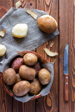 unpeeled and peeled potatoes in a basket and a knife on a wooden background