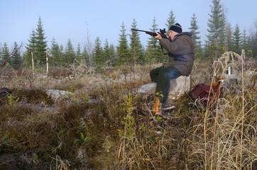 Moose hunter sitting on a stump with a litle fire in front aiming with a rifle, picture from the North of Sweden. 