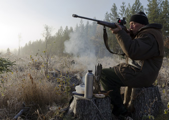 Moose hunter sitting on a stump with a litle smoky fire in front aiming with a rifle and with a...