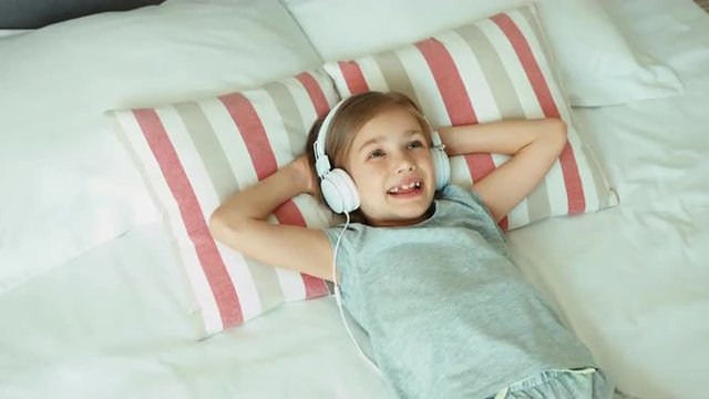 Girl listening music in headphones. Child lying on the bed resting and smiling at camera. Thumb up. Ok